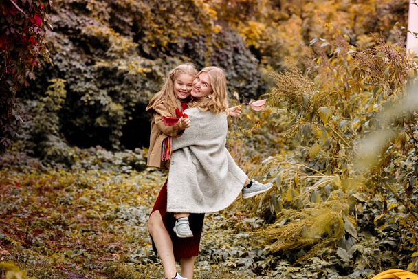 A young adult blonde mother with a 7-year-old daughter on a walk in the autumn park hug and laugh - Фото, зображення