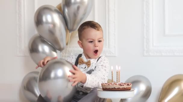 A kisfiú a születésnapját ünnepli, a gyereknek van egy B-napi partija. Gyertyát fúj a szülinapi tortán. Ünnepség, fehér minimalista belső tér. - Felvétel, videó