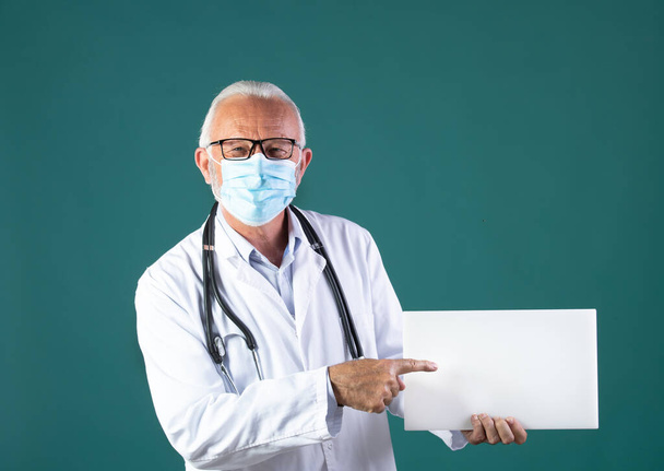 Portrét staršího lékaře v bílém kabátě se stetoskopem kolem krku a ochrannou maskou držící a zobrazující prázdné bílé prkno, na modrém pozadí - Fotografie, Obrázek