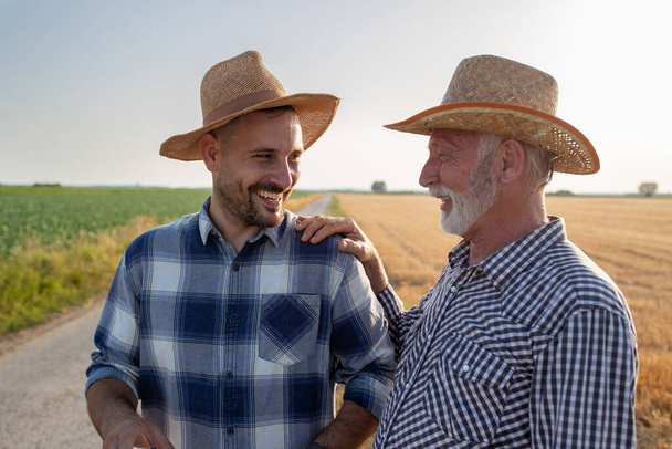 再生シャツとわら帽子をかぶっている2人の農民が話している。夏に畑で収穫中に小麦畑で笑う若い農学者の肩を持つシニア農家.  - 写真・画像