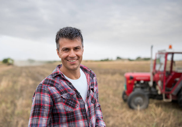 Homem agrônomo olhando para a câmera sorrindo ao usar tablet. Bonito agricultor em pé no campo colhido na frente do trator - Foto, Imagem