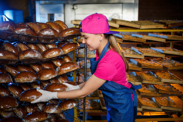 A female baker is at the workplace at the bakery. A professional baker holds bread in his hands. Bread production concept - Zdjęcie, obraz