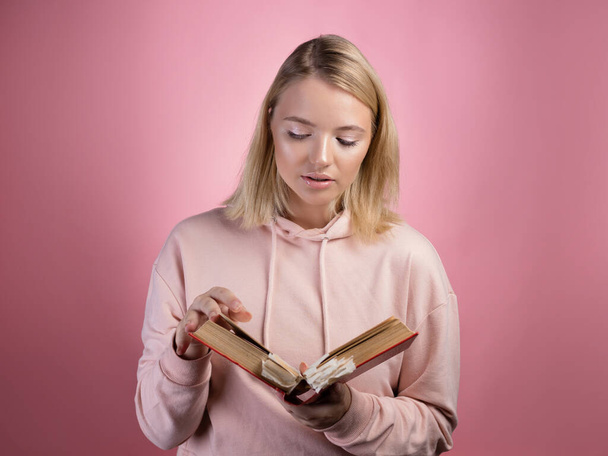 A charming young female student with a book in her hands. A young cute blonde in a pink hoodie - Foto, Imagem