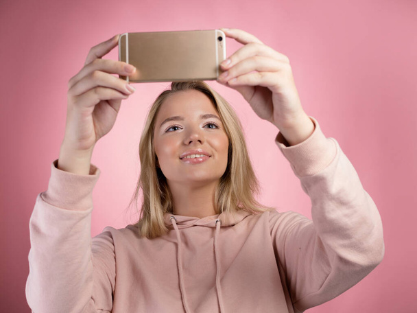 attractive young woman in a pink hoodie uses a smartphone for video communication or photos. - Foto, Imagem