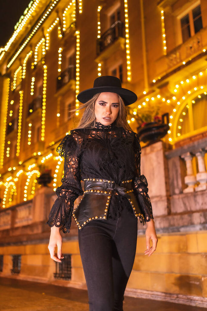 Christmas lights in the city, blonde girl dressed in black with a hat next to an illuminated building in a fashionable pose, winter lifestyle - Fotografie, Obrázek