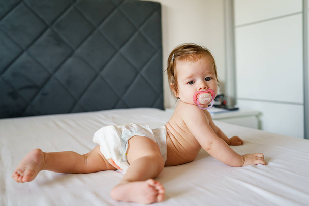 Small caucasian baby girl six months old lying on the bed naked belly down with nipple pacifier wearing diaper at home in bright room copy space - Photo, Image
