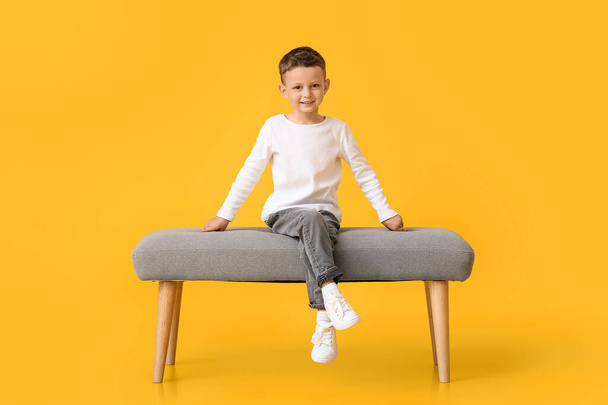 Portrait of cute little boy sitting on bench against color background - Photo, Image
