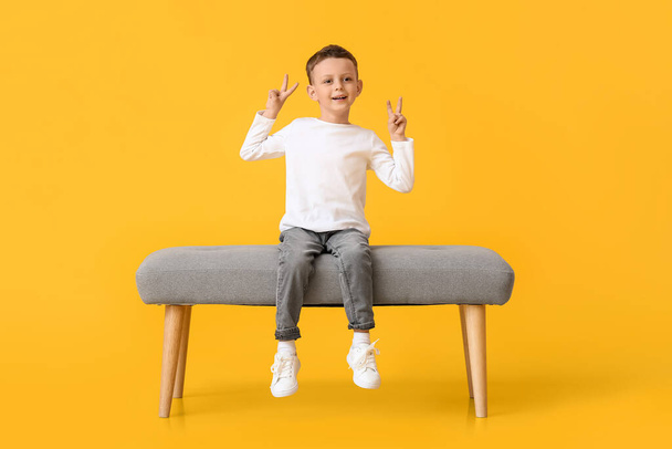 Portrait de mignon petit garçon assis sur le banc et montrant geste de victoire sur fond de couleur - Photo, image