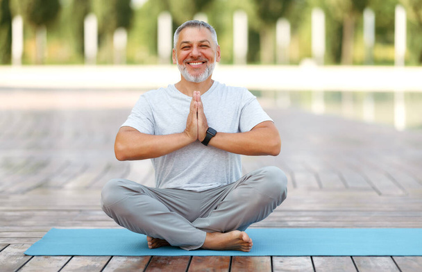 Calm happy mature man sitting in lotus pose on mat during morning meditation in park - Photo, image