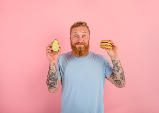 Glücklicher Mann mit Bart und Tattoos ist unentschlossen, ob er eine Avocado oder einen Hamburger essen soll - Foto, Bild