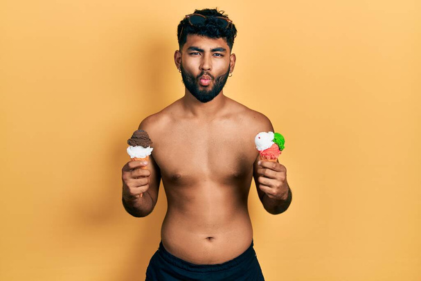 Arabischer Mann mit Bart in Badebekleidung, der zwei Eistüten isst und Fischgesicht mit Mund und zusammengekniffenen Augen macht, verrückt und komisch.  - Foto, Bild