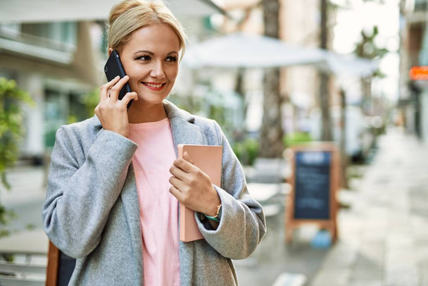 Νεαρή ξανθιά επιχειρηματίας χαμογελώντας ευτυχισμένη μιλώντας στο smartphone στην πόλη. - Φωτογραφία, εικόνα