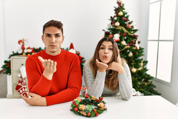 Ein junges hispanisches Paar, das an Weihnachten am Tisch sitzt und in die Kamera schaut, bläst einen Kuss mit der Hand in die Luft, der schön und sexy ist. Liebesausdruck.  - Foto, Bild