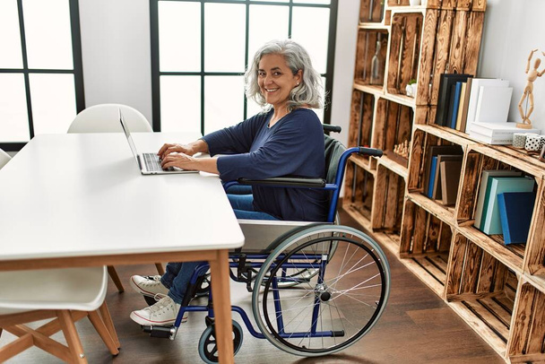 Keski-ikäinen harmaa tukka vammainen nainen, joka istuu kotona pyörätuolilla kannettavan tietokoneen avulla. - Valokuva, kuva