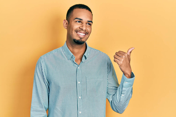 Молодой африканский американец в повседневной одежде улыбается со счастливым лицом, глядя и указывая на сторону с большим пальцем вверх.  - Фото, изображение