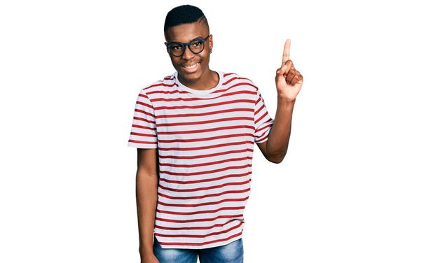 Młody Afroamerykanin w luźnej koszuli i okularach pokazuje i wskazuje palcem numer jeden, uśmiechając się pewny siebie i szczęśliwy..  - Zdjęcie, obraz