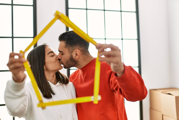 Jovem casal latino beijando e segurando casa projeto nova casa. - Foto, Imagem