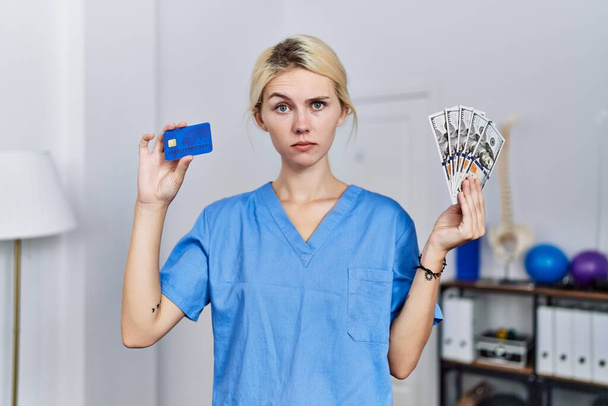 Молодая физиотерапевт женщина с кредиткой и долларами банкноты скептически и нервно, хмурясь расстроен из-за проблемы. отрицательный человек.  - Фото, изображение