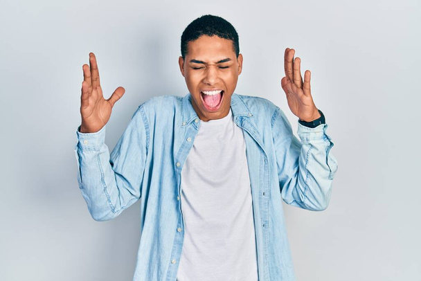 Νεαρός Αφροαμερικάνος που φοράει καθημερινά ρούχα γιορτάζει τρελός και τρελός για την επιτυχία με τα χέρια σηκωμένα και τα κλειστά μάτια να ουρλιάζουν ενθουσιασμένοι. έννοια νικητή  - Φωτογραφία, εικόνα