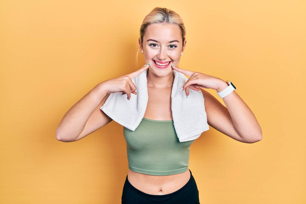 Молодая блондинка в спортивной одежде и полотенце улыбается с открытым ртом, пальцы указывают и заставляя веселую улыбку  - Фото, изображение