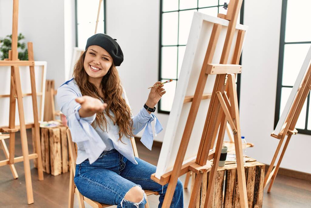 Młoda latynoska artystka malująca na płótnie w pracowni sztuki uśmiechnięta, przyjazna, oferująca uścisk dłoni jako powitanie i powitanie. udany biznes.  - Zdjęcie, obraz