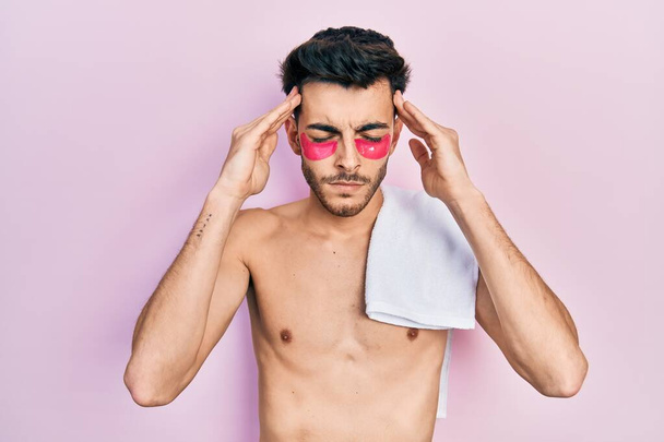 Jonge Spaanse man shirtloos het dragen van handdoek en oogzakken pleisters die lijden aan hoofdpijn wanhopig en gestresst omdat pijn en migraine. handen op het hoofd.  - Foto, afbeelding