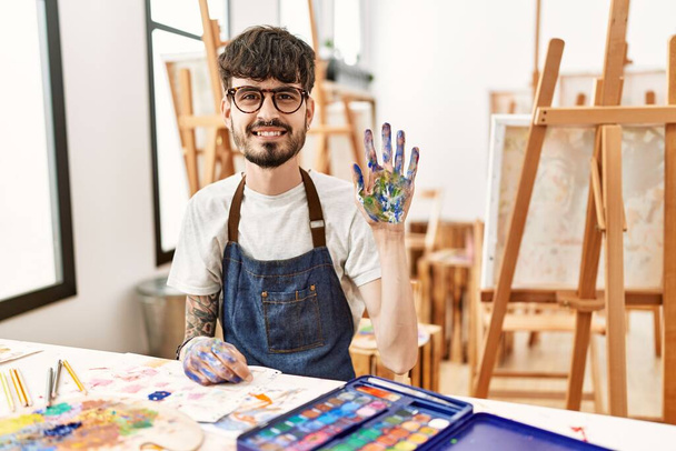 Spanyol férfi szakállal a művészeti stúdióban bemutatja és felmutat az ötös számú ujjaival, miközben magabiztosan és boldogan mosolyog..  - Fotó, kép
