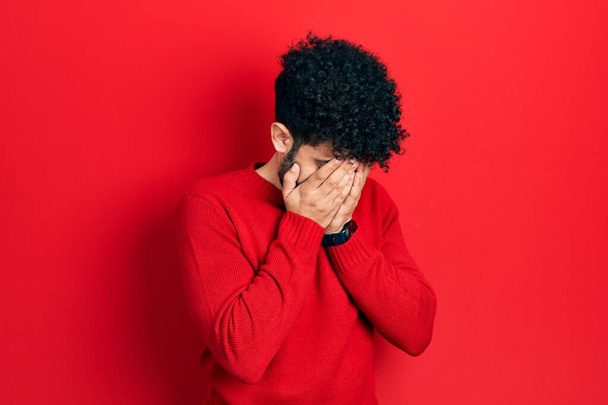 Mladý arabský muž s vousy na sobě ležérní červený svetr se smutným výrazem zakrývající tvář s rukama při pláči. deprese koncept.  - Fotografie, Obrázek