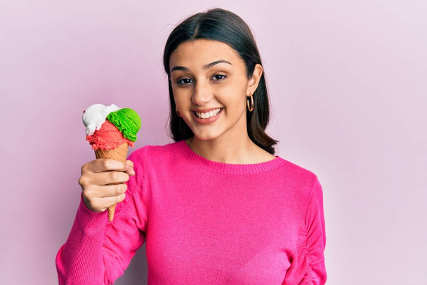 Young hispanic woman holding ice cream looking positive and happy standing and smiling with a confident smile showing teeth  - Фото, зображення