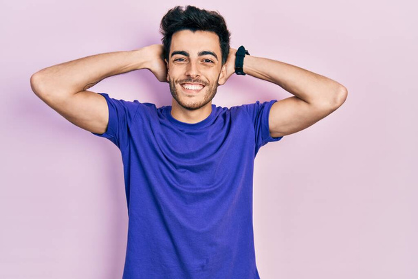 Fiatal spanyol férfi alkalmi pólót visel pihentető és nyújtózkodó, karok és kezek a fej és a nyak mögött mosolyogva boldog  - Fotó, kép