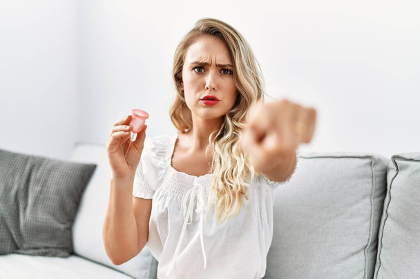 Mujer joven rubia sosteniendo la copa menstrual señalando con el dedo a la cámara y a usted, gesto de confianza mirando en serio  - Foto, Imagen