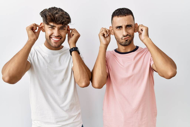 Young gay couple standing over isolated background smiling pulling ears with fingers, funny gesture. audition problem  - Photo, Image