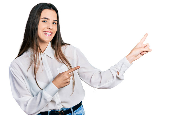 Νεαρή μελαχρινή έφηβη που φοράει επαγγελματική λευκή μπλούζα χαμογελώντας και κοιτάζοντας την κάμερα που δείχνει με τα δύο χέρια και τα δάχτυλα στο πλάι.  - Φωτογραφία, εικόνα