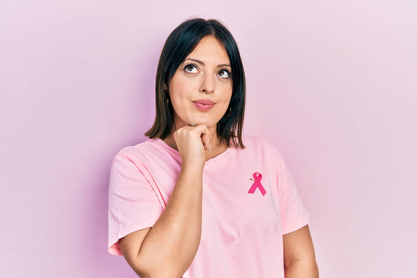 若いヒスパニック系の女性は、質問について考えてあごの手でTシャツの上にピンクの癌のリボンを着て、集中的な表現。笑顔で思いやりのある顔。疑わしい概念.  - 写真・画像
