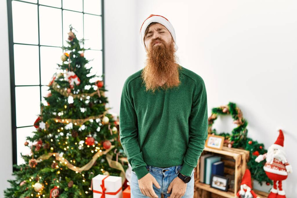 Pelirrojo con barba larga usando sombrero de navidad por árbol de navidad mirando a la cámara soplando un beso en el aire siendo encantador y sexy. expresión de amor.  - Foto, imagen
