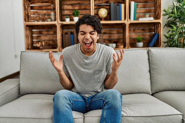 Mladý hispánec, oblečený do neformálního oblečení, sedí doma na pohovce a oslavuje šílený a šílený úspěch se zdviženýma rukama a zavřenýma očima a vzrušeně křičí. koncept vítěze  - Fotografie, Obrázek