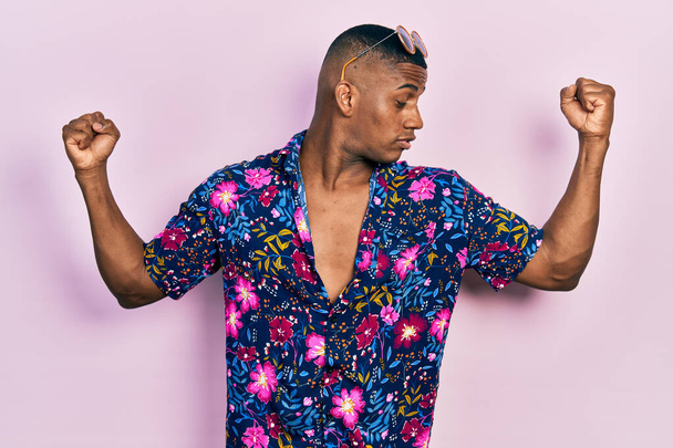 Young black man wearing hawaiian shirt and sunglasses showing arms muscles smiling proud. fitness concept.  - Foto, Bild
