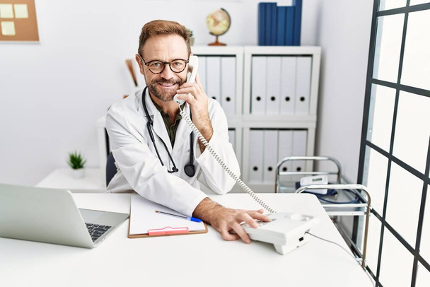 Middle age hispanic man wearing doctor uniform talking on the telephone at clinic - Fotografie, Obrázek