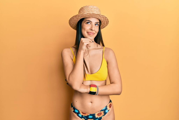 質問について考える顎の手でビキニと夏の帽子を身に着けている若いヒスパニック系の女性,集中的な表現.思慮深い顔で笑顔。疑わしい概念.  - 写真・画像