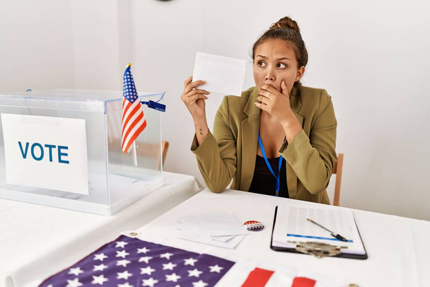 Beautiful hispanic woman holding voting envelop in ballot box covering mouth with hand, shocked and afraid for mistake. surprised expression  - Photo, Image