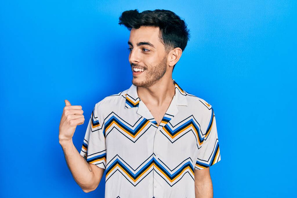 Jeune homme hispanique portant des vêtements décontractés souriant avec le visage heureux regardant et pointant vers le côté avec le pouce vers le haut.  - Photo, image