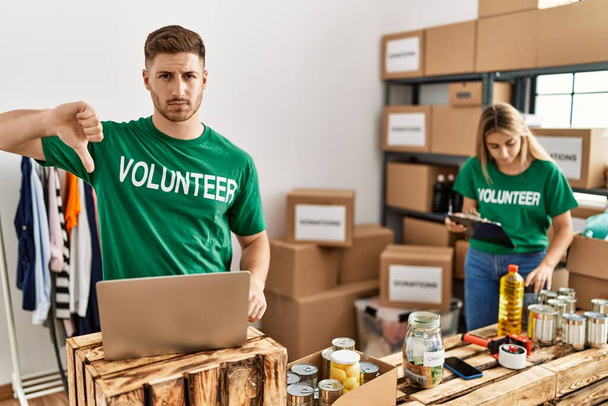 Young man and woman wearing volunteer t shirt at donations stand with angry face, negative sign showing dislike with thumbs down, rejection concept  - 写真・画像
