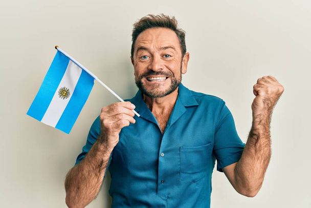 Mann mittleren Alters mit Argentinien-Fahne schreit stolz, feiert Sieg und Erfolg sehr aufgeregt mit erhobenem Arm  - Foto, Bild