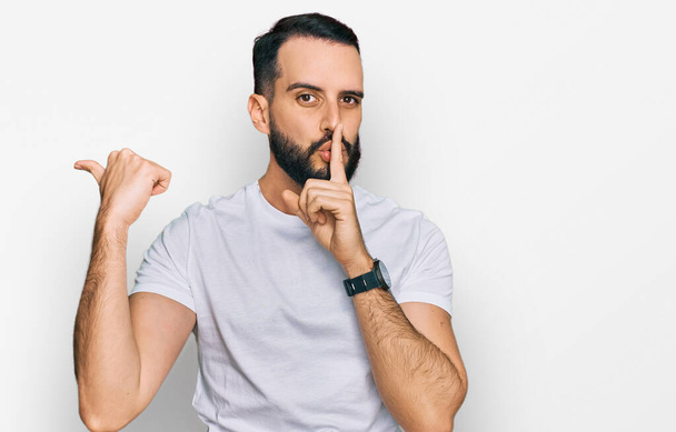 Young man with beard wearing casual white t shirt asking to be quiet with finger on lips pointing with hand to the side. silence and secret concept.  - Φωτογραφία, εικόνα