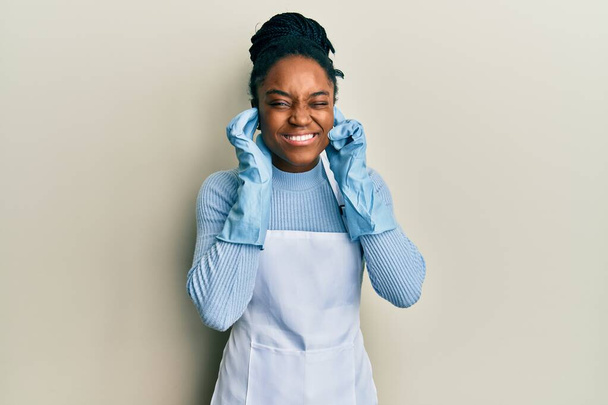 Африканская американка с плетеными волосами в чистом фартуке и перчатках, покрывающих уши пальцами с раздраженным выражением для шума громкой музыки. Концепция deaf.  - Фото, изображение