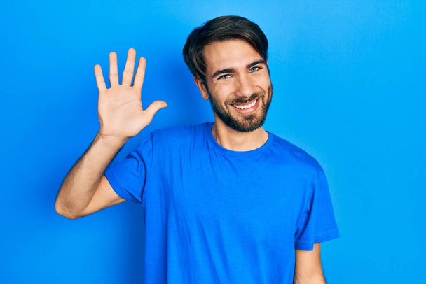 Joven hombre hispano usando ropa casual renunciando a decir hola feliz y sonriente, gesto de bienvenida amistoso  - Foto, imagen