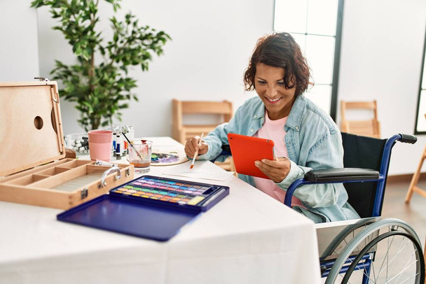 Mujer artista discapacitada hispana de mediana edad dibujando usando touchpad sentado en silla de ruedas en el estudio de arte. - Foto, Imagen