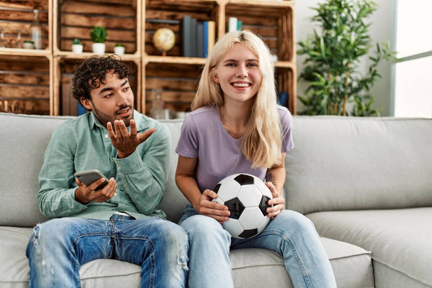 Nainen hymyilee onnellinen katsomassa jalkapallo-ottelua ja poikaystävä tylsä älypuhelimella kotona. - Valokuva, kuva