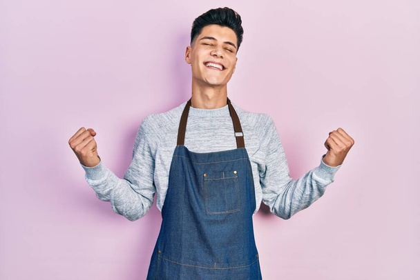 Young hispanic man wearing apron very happy and excited doing winner gesture with arms raised, smiling and screaming for success. celebration concept.  - Photo, Image