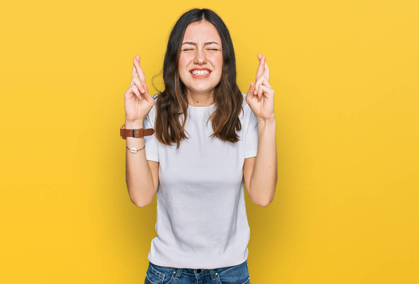 Young beautiful woman wearing casual white t shirt gesturing finger crossed smiling with hope and eyes closed. luck and superstitious concept.  - Foto, imagen